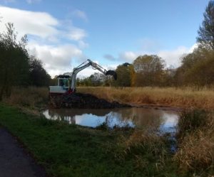 Callander Froglife - mechanical digger excavating near edge of pond. Reflections of blue sky and trees in pond surface.