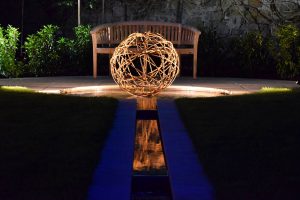 Garden Lighting, garden water feature, floating metal ball structure lit up at night by hidden lighting, wooden bench seat in background